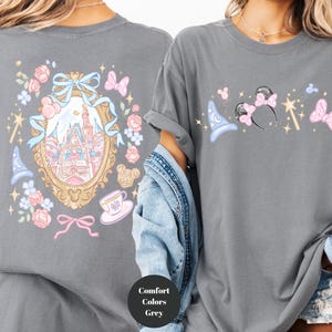 May include: A gray t-shirt featuring a whimsical design. The front displays a collection of magical icons, including a wizard hat, mouse ears, a wand, and butterflies. The back showcases a castle within an ornate frame, surrounded by floral and bow accents.