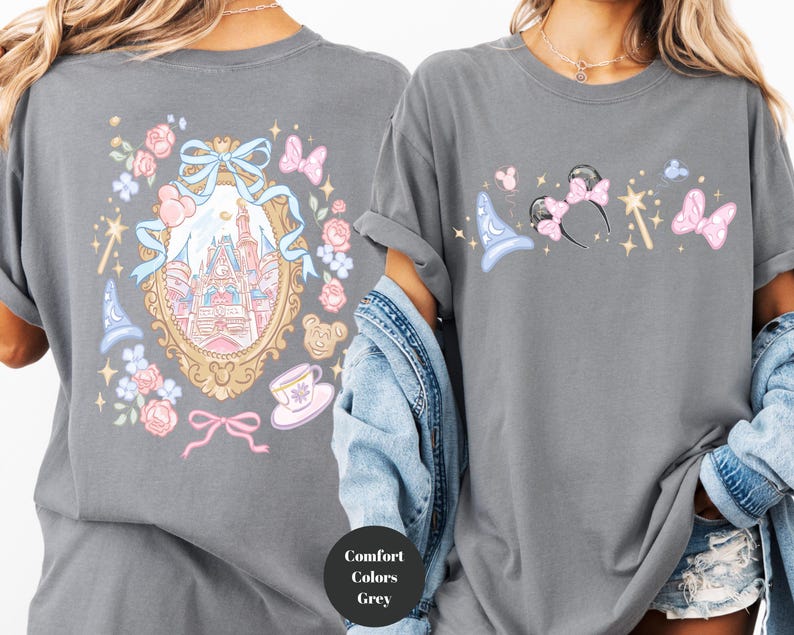 May include: A gray t-shirt featuring a whimsical design. The front displays a collection of magical icons, including a wizard hat, mouse ears, a wand, and butterflies. The back showcases a castle within an ornate frame, surrounded by floral and bow accents.