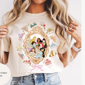 May include: Ivory-colored t-shirt featuring a gold-framed portrait of Disney princesses. Surrounding the frame are various princess-themed crowns and accessories. The shirt has the words "Comfort Colors Ivory" printed on it.