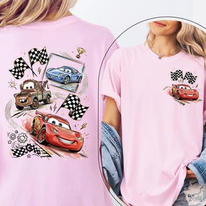 Puede incluir: Una camiseta rosa claro con coches de carreras de dibujos animados y banderas a cuadros. El dise&ntilde;o incluye un coche de carreras rojo, un coche azul y una gr&uacute;a marr&oacute;n. Un peque&ntilde;o dise&ntilde;o de un coche de carreras rojo y banderas a cuadros est&aacute; en el bolsillo.