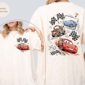 Puede incluir: Camiseta de color marfil con un gr&aacute;fico de la pel&iacute;cula Cars. La parte delantera muestra a Rayo McQueen con banderas a cuadros. La parte trasera muestra a Rayo McQueen, Mate y un coche azul, junto con banderas a cuadros y un trofeo.
