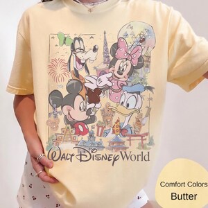 May include: A butter yellow t-shirt featuring a graphic print of Mickey Mouse, Minnie Mouse, Goofy, and Donald Duck in front of Walt Disney World landmarks. The text Walt Disney World is printed in a dark script font below the colorful character illustration.