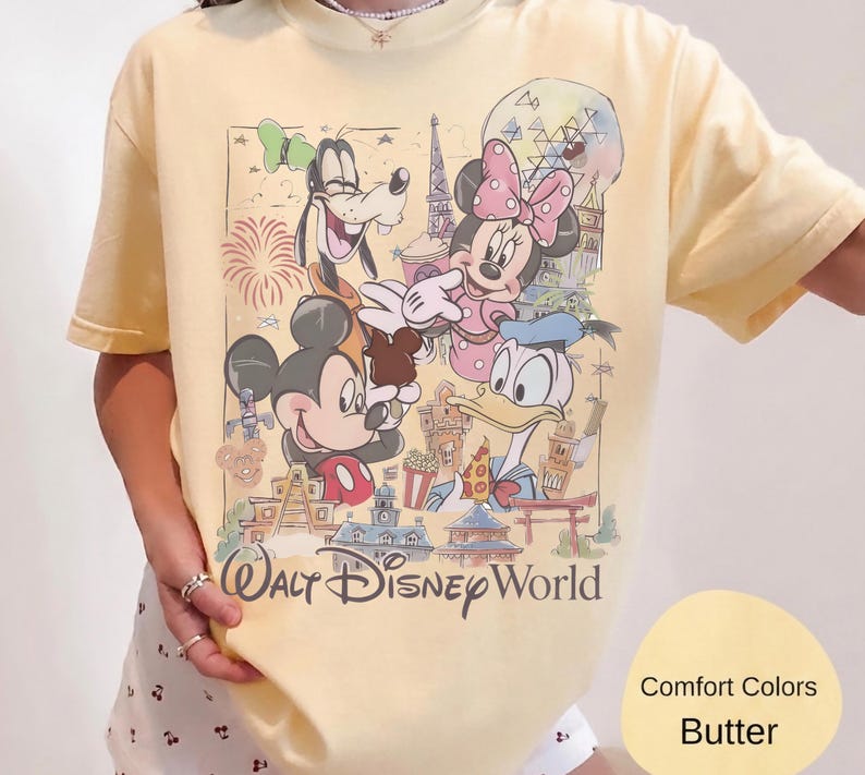 May include: A butter yellow t-shirt featuring a graphic print of Mickey Mouse, Minnie Mouse, Goofy, and Donald Duck in front of Walt Disney World landmarks. The text Walt Disney World is printed in a dark script font below the colorful character illustration.