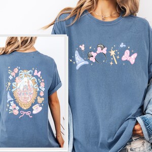 May include: A blue t-shirt featuring a whimsical design with a castle, a magic wand, and mouse ears. The back of the shirt has a framed castle design with floral and bow accents. The shirt is made of a soft material.