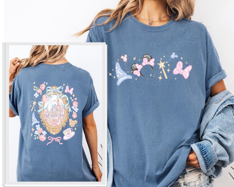 May include: A blue t-shirt featuring a whimsical design with a castle, a magic wand, and mouse ears. The back of the shirt has a framed castle design with floral and bow accents. The shirt is made of a soft material.