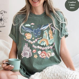 K&ouml;nnte beinhalten: Ein gr&uuml;nes T-Shirt mit einem grafischen Druck eines Drachen, eines Schlosses, eines Regenbogens und Figuren aus einem beliebten Videospiel. Der Text "EPIC UNIVERSE" ist auf dem Shirt gedruckt.