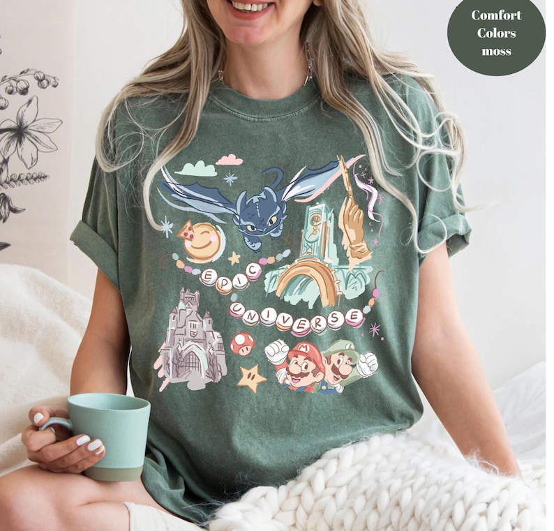 K&ouml;nnte beinhalten: Ein gr&uuml;nes T-Shirt mit einem grafischen Druck eines Drachen, eines Schlosses, eines Regenbogens und Figuren aus einem beliebten Videospiel. Der Text "EPIC UNIVERSE" ist auf dem Shirt gedruckt.