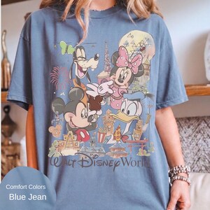 May include: A blue jean colored short-sleeve t-shirt featuring a graphic print of Mickey Mouse, Minnie Mouse, Goofy, and Donald Duck. The design includes various park landmarks, fireworks, and the text Walt Disney World printed at the bottom.