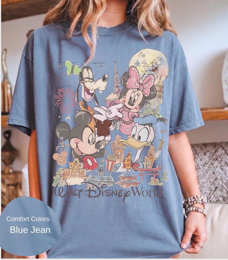 May include: A blue jean colored short-sleeve t-shirt featuring a graphic print of Mickey Mouse, Minnie Mouse, Goofy, and Donald Duck. The design includes various park landmarks, fireworks, and the text Walt Disney World printed at the bottom.