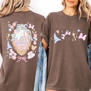 May include: Brown t-shirts with Disney-themed graphics. One shirt features a castle design within a gold frame, surrounded by flowers, bows, and a teacup. The other shirt displays a wizard hat, Minnie Mouse ears, a wand, and a bow.
