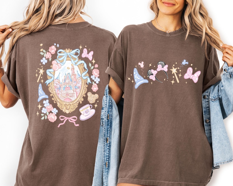May include: Brown t-shirts with Disney-themed graphics. One shirt features a castle design within a gold frame, surrounded by flowers, bows, and a teacup. The other shirt displays a wizard hat, Minnie Mouse ears, a wand, and a bow.