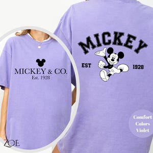 Puede incluir: Una camiseta morada con un gr&aacute;fico de Mickey Mouse negro y el texto "Mickey & Co. Est. 1928" en la parte delantera. La parte posterior de la camiseta tiene un gr&aacute;fico de Mickey Mouse m&aacute;s grande con el texto "Mickey Est. 1928".
