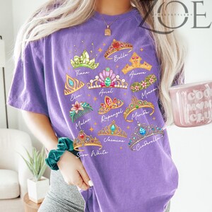 K&ouml;nnte beinhalten: Ein lila T-Shirt mit farbenfrohen Illustrationen von Prinzessinnenkronen mit Namen wie Belle, Elsa und Moana. Das Shirt hat einen Rundhalsausschnitt und kurze &Auml;rmel. Der Text "ZOE THE STYLEERY" ist oben rechts aufgedruckt.