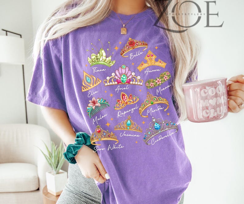 K&ouml;nnte beinhalten: Ein lila T-Shirt mit farbenfrohen Illustrationen von Prinzessinnenkronen mit Namen wie Belle, Elsa und Moana. Das Shirt hat einen Rundhalsausschnitt und kurze &Auml;rmel. Der Text "ZOE THE STYLEERY" ist oben rechts aufgedruckt.