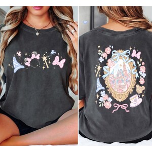 May include: Dark gray t-shirt featuring a whimsical design with a magic wand, Minnie Mouse ears, butterflies, and a wizard hat on the front. The back showcases a castle within a decorative frame, surrounded by flowers and bows.