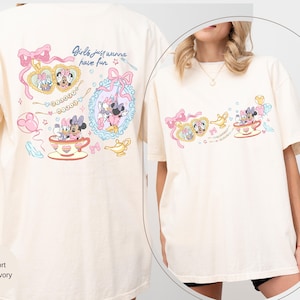 May include: Cream-coloured t-shirt with a vibrant graphic of Minnie Mouse and Daisy Duck. The design includes the text "Girls just wanna have fun" and decorative elements such as bows, teacups, and jewellery. The shirt has short sleeves and a relaxed fit.