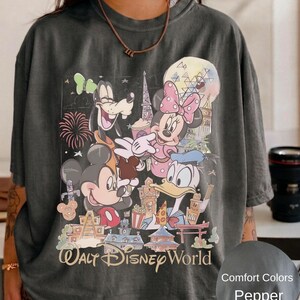 May include: A charcoal gray t-shirt featuring a graphic print of Mickey Mouse, Minnie Mouse, Goofy, and Donald Duck in front of various park landmarks. The text Walt Disney World is printed at the bottom in a tan script font. A small circular swatch shows the color label Comfort Colors Pepper.
