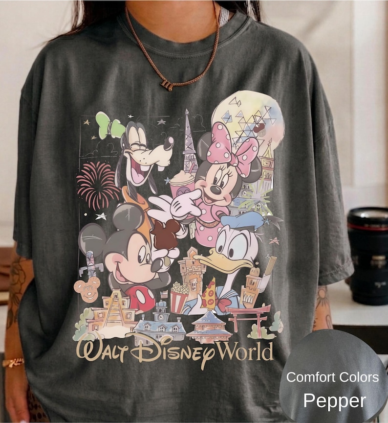 May include: A charcoal gray t-shirt featuring a graphic print of Mickey Mouse, Minnie Mouse, Goofy, and Donald Duck in front of various park landmarks. The text Walt Disney World is printed at the bottom in a tan script font. A small circular swatch shows the color label Comfort Colors Pepper.