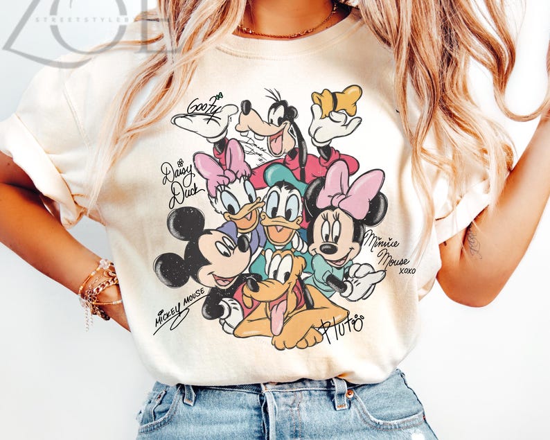 May include: Cream-colored t-shirt featuring a graphic of Disney characters including Mickey Mouse, Minnie Mouse, Donald Duck, Daisy Duck, Goofy, and Pluto. The characters are surrounded by their signatures. The shirt is a casual wear item.