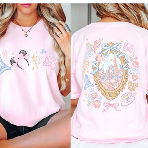 May include: A light pink t-shirt featuring a front design with a blue wizard hat, Minnie Mouse ears, a wand, and a pink bow. The back of the shirt has a castle design inside a gold frame, surrounded by flowers, bows, and a teacup.