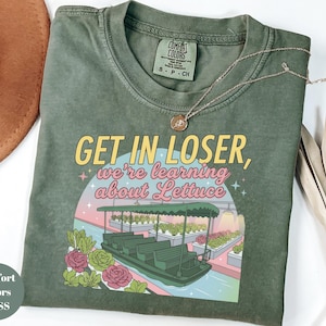 Puede incluir: Una camiseta de manga corta color verde musgo con un estampado gr&aacute;fico que muestra un barco en un invernadero y el texto "GET IN LOSER, we're learning about Lettuce". La camiseta tiene cuello redondo y una etiqueta Comfort Colors en el interior.