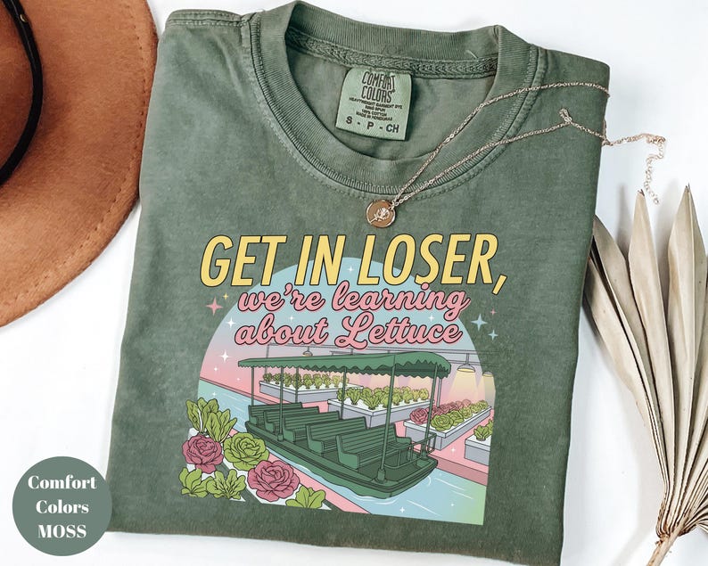 Puede incluir: Una camiseta de manga corta color verde musgo con un estampado gr&aacute;fico que muestra un barco en un invernadero y el texto "GET IN LOSER, we're learning about Lettuce". La camiseta tiene cuello redondo y una etiqueta Comfort Colors en el interior.