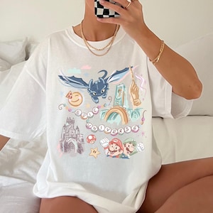 K&ouml;nnte beinhalten: Wei&szlig;es T-Shirt mit einem bunten Grafikdruck, der einen Drachen, ein Schloss und Figuren aus dem Videospiel Super Mario Bros. zeigt. Der Text "Epic Universe" ist auf dem Shirt gedruckt.