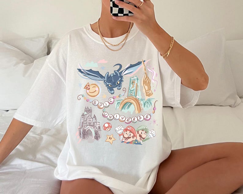 K&ouml;nnte beinhalten: Wei&szlig;es T-Shirt mit einem bunten Grafikdruck, der einen Drachen, ein Schloss und Figuren aus dem Videospiel Super Mario Bros. zeigt. Der Text "Epic Universe" ist auf dem Shirt gedruckt.