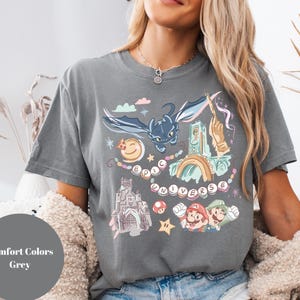 K&ouml;nnte beinhalten: Ein graues T-Shirt mit einem grafischen Druck eines Drachen, eines Schlosses und Figuren aus einem beliebten Videospiel. Der Text "Epic Universe" ist auf dem Shirt gedruckt.