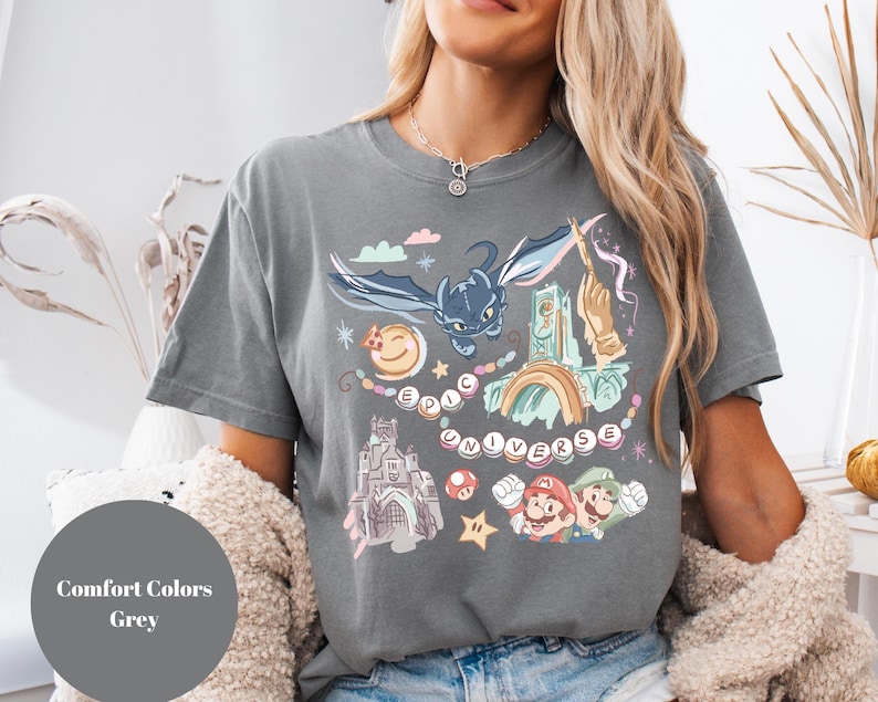 K&ouml;nnte beinhalten: Ein graues T-Shirt mit einem grafischen Druck eines Drachen, eines Schlosses und Figuren aus einem beliebten Videospiel. Der Text "Epic Universe" ist auf dem Shirt gedruckt.