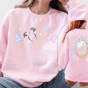 May include: A light pink sweatshirt featuring whimsical illustrations. The front displays a blue wizard hat, Minnie Mouse ears, a magic wand, and a pink bow. The back showcases a castle design within a decorative frame.