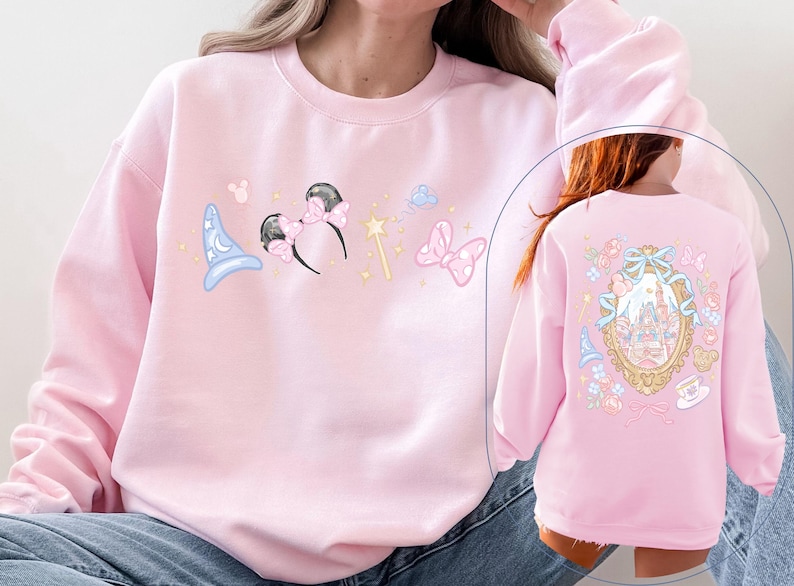 May include: A light pink sweatshirt featuring whimsical illustrations. The front displays a blue wizard hat, Minnie Mouse ears, a magic wand, and a pink bow. The back showcases a castle design within a decorative frame.