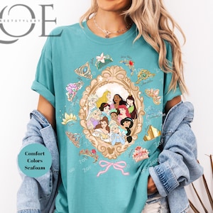 May include: Seafoam green t-shirt featuring a framed illustration of Disney princesses. The frame is surrounded by decorative elements and a pink bow. The shirt has the text "Comfort Colors Seafoam". The brand name "Zoe Street Style" is at the top left.