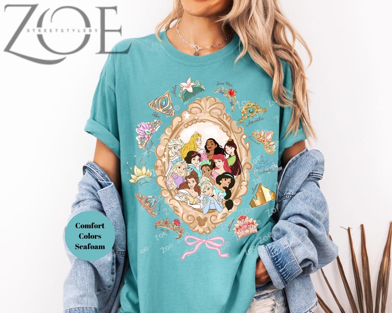 May include: Seafoam green t-shirt featuring a framed illustration of Disney princesses. The frame is surrounded by decorative elements and a pink bow. The shirt has the text "Comfort Colors Seafoam". The brand name "Zoe Street Style" is at the top left.