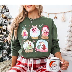 May include: Green sweatshirt featuring six snow globes with princess illustrations. The snow globes have pink bows, candy canes, and other festive details. A Santa mug is held in the hand.