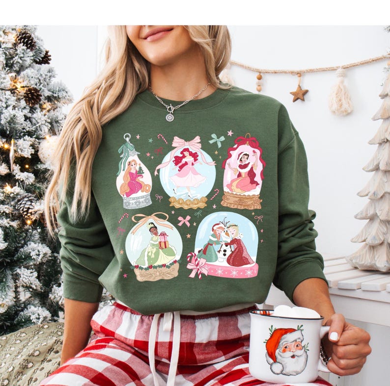 May include: Green sweatshirt featuring six snow globes with princess illustrations. The snow globes have pink bows, candy canes, and other festive details. A Santa mug is held in the hand.