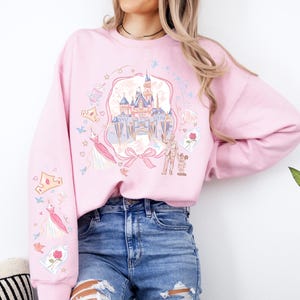 May include: Pink sweatshirt with a castle graphic, princess dress, and crown illustrations. The design includes a castle, a bow, and characters. The sleeves have additional graphics of a crown, dress, and rose.
