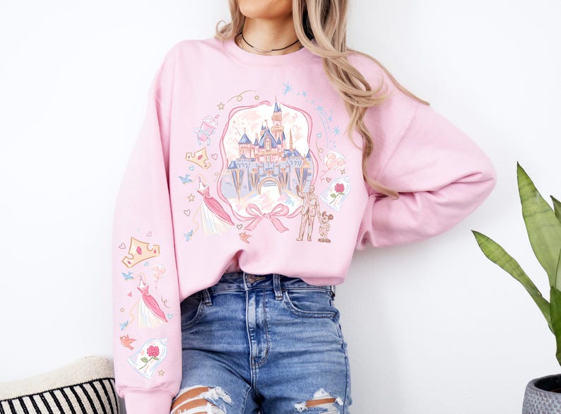 May include: Pink sweatshirt with a castle graphic, princess dress, and crown illustrations. The design includes a castle, a bow, and characters. The sleeves have additional graphics of a crown, dress, and rose.