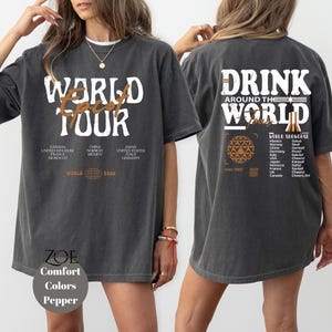 May include: A dark gray oversized t-shirt with a white graphic print that says "World Tour" on the front and "Drink Around the World" on the back. The back graphic also includes a list of countries and a world map.