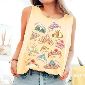 May include: Yellow tank top featuring illustrations of various princess tiaras with names like Ariel, Belle, and Cinderella. The design includes colorful crowns with gems and floral accents, perfect for fans of fairy tales.