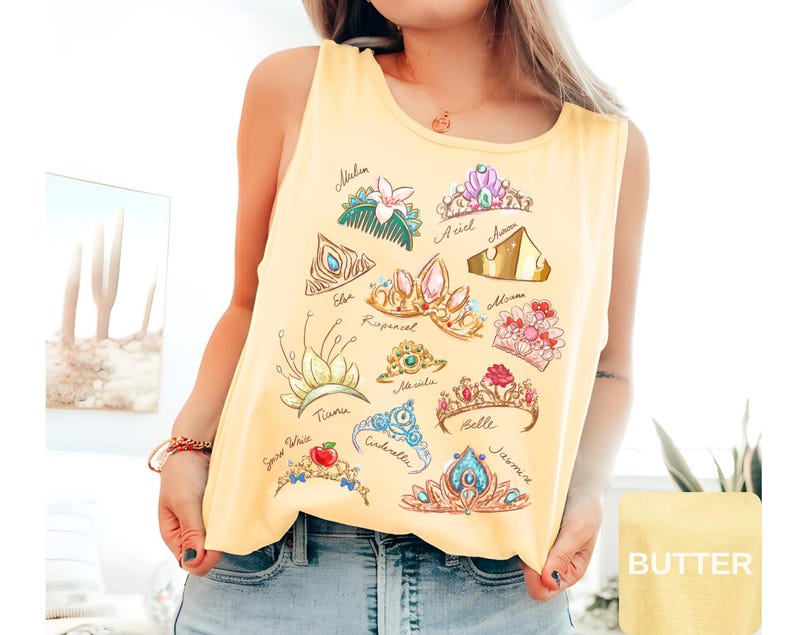 May include: Yellow tank top featuring illustrations of various princess tiaras with names like Ariel, Belle, and Cinderella. The design includes colorful crowns with gems and floral accents, perfect for fans of fairy tales.