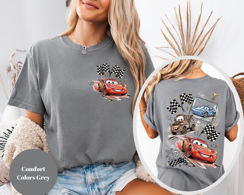 Puede incluir: Camiseta gris con un gr&aacute;fico de Rayo McQueen de la pel&iacute;cula Cars, con banderas a cuadros. La parte posterior de la camiseta muestra a Rayo McQueen, Mate y un coche azul. El texto "Comfort Colors Grey" es visible.