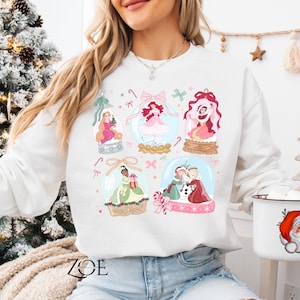 May include: White sweatshirt featuring colorful illustrations of princesses inside snow globes. The design includes pink bows, candy canes, and decorative elements. A Santa mug is held in the hand.