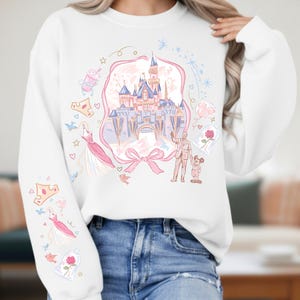 May include: White sweatshirt featuring a castle illustration with pink and blue accents. The design includes a pink ribbon, a dress, a rose, and other whimsical elements. The sleeves have additional illustrations of crowns and dresses.