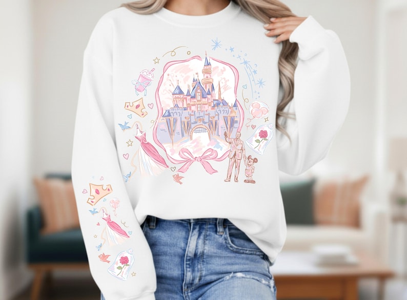 May include: White sweatshirt featuring a castle illustration with pink and blue accents. The design includes a pink ribbon, a dress, a rose, and other whimsical elements. The sleeves have additional illustrations of crowns and dresses.