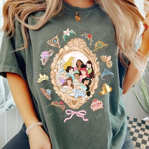 May include: Dark green t-shirt featuring a framed illustration of various cartoon princesses. The frame is surrounded by tiaras and floral accents. A pink bow is at the bottom of the frame. The text "Zoe" is repeated around the design.