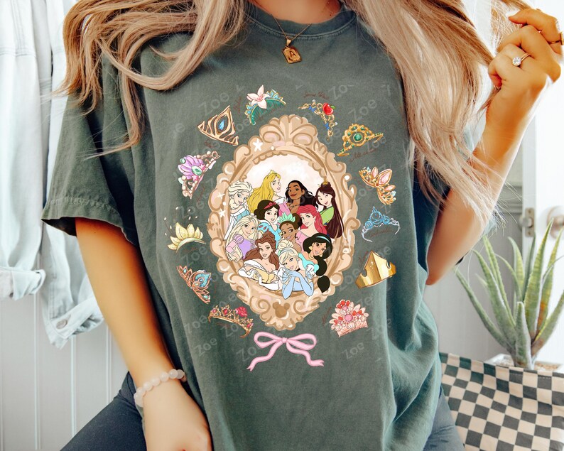 May include: Dark green t-shirt featuring a framed illustration of various cartoon princesses. The frame is surrounded by tiaras and floral accents. A pink bow is at the bottom of the frame. The text "Zoe" is repeated around the design.