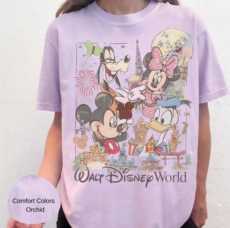 May include: A light purple t-shirt featuring a graphic print of Mickey Mouse, Minnie Mouse, Goofy, and Donald Duck. The design includes various park landmarks, fireworks, and the text Walt Disney World. The shirt is labeled as Comfort Colors Orchid.