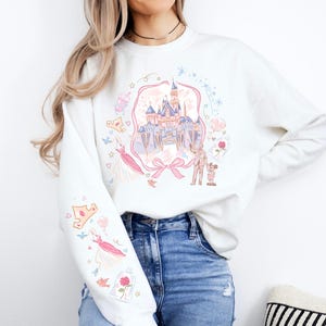 May include: White sweatshirt featuring a colorful illustration of a castle, characters, and other elements. The design includes pink, blue, and yellow accents. The sleeve has additional graphics. A bow is at the bottom of the castle.