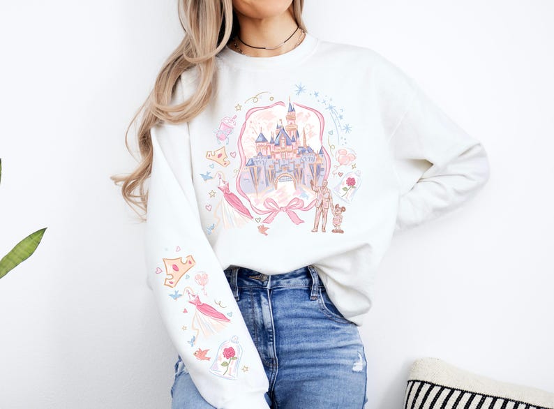 May include: White sweatshirt featuring a colorful illustration of a castle, characters, and other elements. The design includes pink, blue, and yellow accents. The sleeve has additional graphics. A bow is at the bottom of the castle.